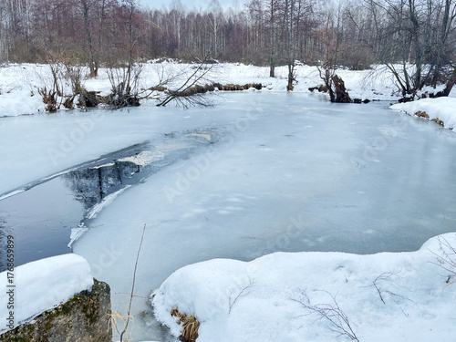Scenic winter riverbank with snow-covered terrain and partially frozen water, reflecting the surrounding trees and creating a peaceful, serene atmosphere