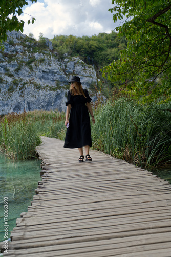 A girl in black walks peacefully along a wooden boardwalk at Plitvice Lake, surrounded by lush reeds and rocky cliffs under a partly cloudy sky—serene nature embracing quiet solitude.