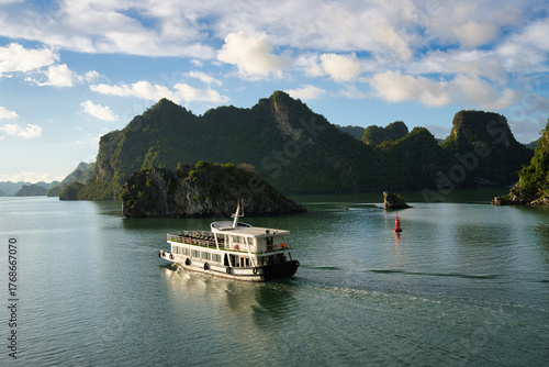 Tourist boat sailing on the calm sea of ha long bay, with towering limestone karsts emerging from the water under clear blue sky, creating a famous travel destination