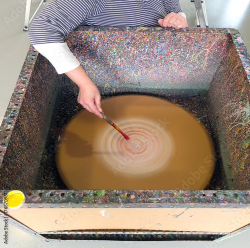 Konzept Kunst, Unterricht und Kunstpädagogik: Draufsicht auf eine Frau, die Farbtropfen von einem Pinsel auf eine rotierende Scheibe in einer Box mit Farbspritzern fallen lässt