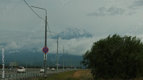 View of majestic snow-capped volcano rising above clouds and heavy traffic on highway at its foot.
