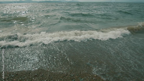 Waves crash onto shore creating foam at edges and washing pebbles out of sand. Water's surface sparkles in sun and hazy mountain range is visible on horizon. Slow motion