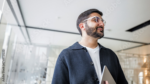 Fotografía Man stands in a modern office holding a laptop and looking thoughtfully towards