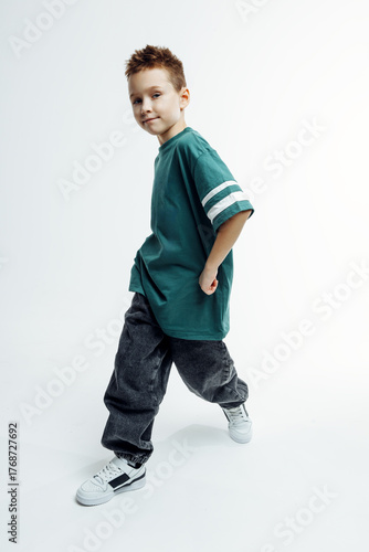 Young boy with short hair wearing a teal oversized t-shirt and baggy jeans, posing confidently with a playful expression against a bright, minimalistic background