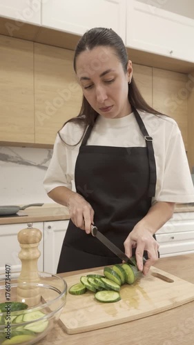 Young woman in black apron slicing cucumber on wooden board in bright kitchen, preparing fresh healthy meal in calm domestic atmosphere with natural daylight