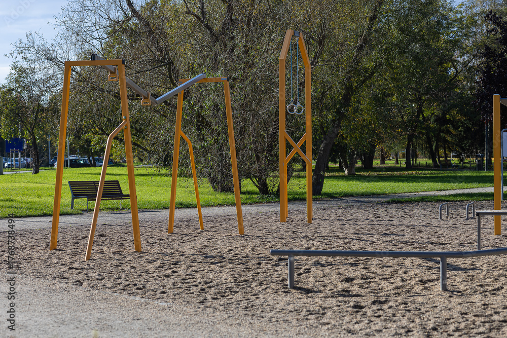 Fototapeta premium Playground equipment featuring modern swings and gymnastic rings, set in a vibrant park with lush greenery and a sandy surface, inviting outdoor play and fitness activities for children and adults