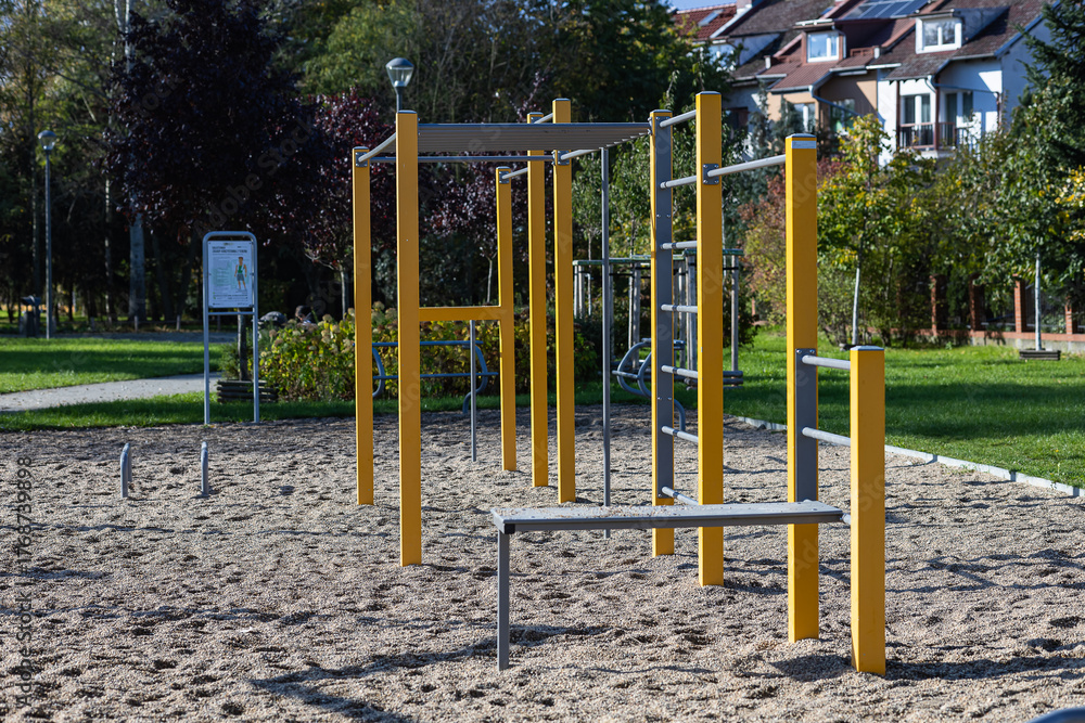 Fototapeta premium Outdoor playground featuring yellow exercise bars and sandy ground, surrounded by green grass and trees, providing a vibrant space for physical activity and recreation in a community park setting