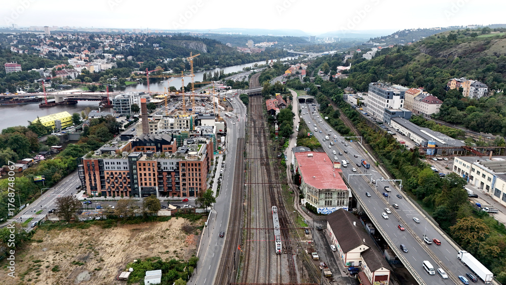 Fototapeta premium Aerial view of cityscape with parallel railway tracks and busy highway beside river. Urban contrast between movement, architecture, and industrial landscape captured from above.