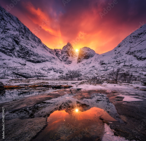 Vibrant orange and red sunrise illuminating snow-covered peaks, reflecting on the icy surface of frozen lake in a tranquil winter mountain landscape. Snowy mountains and frozen fjord. Lofoten, Norway