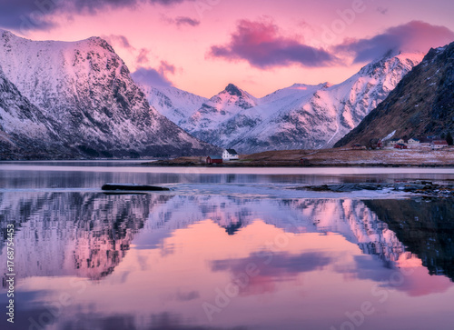 Beautiful pink sunrise illuminating snowy mountains and houses reflecting in the calm water of a fjord in Lofoten islands, Norway. Winter landscape with sea, pink sky with clouds, rorbu, rocks in snow