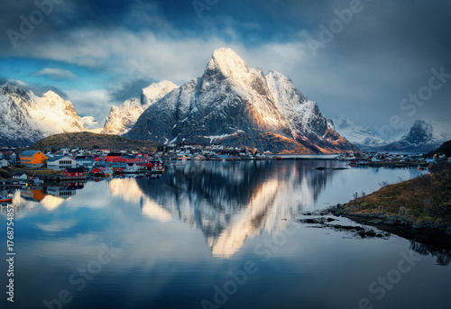 Snowy mountains and colorful houses reflect in a fjord under dramatic clouds at sunset. Winter scenery with sea, rocks in snow, cloudy blue sky, rorbu. Reine village in Lofoten, Norway. Landscape