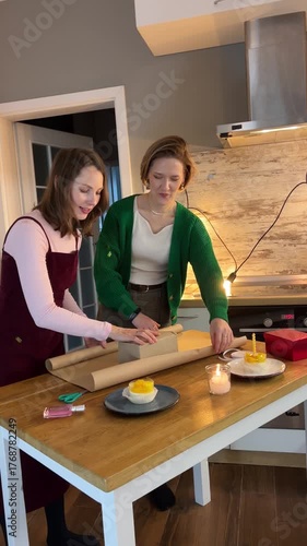 Friends preparing present at kitchen island under Happy Birthday garland and string lights, cozy indoor celebration setup.