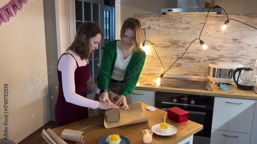Adult women arranging birthday gift wrap in modern kitchen decorated for party, teamwork and cheerful home celebration mood