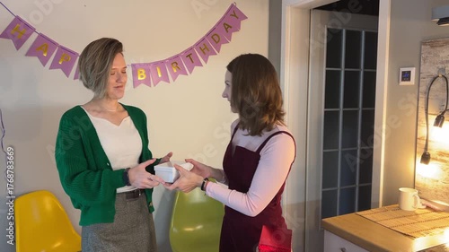 Woman giving wrapped birthday present to friend in decorated kitchen under 