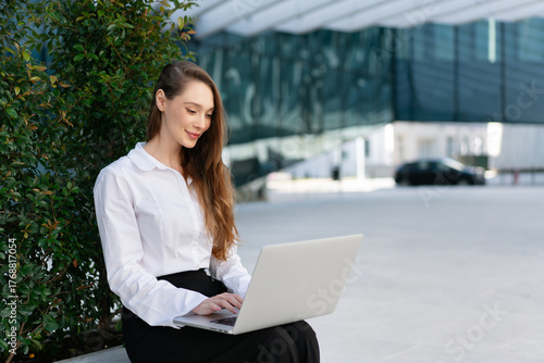 Smiling young businesswoman using laptop while working on project. Happy young woman in white shirt sitting near plants with blurred office building in background