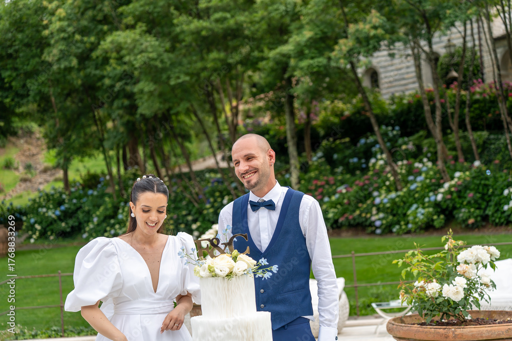 Fototapeta premium Joyful couple celebrating by cutting the wedding cake surrounded by friends and family