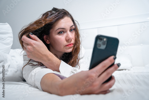 A woman lies on a bed in a white room using a smartphone to take a selfie. she casually fixes her hair, capturing a moment of relaxation and self-expression.