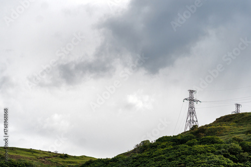 Powerline towers on green hillside beneath stormy sky. Electric transmission towers rise above lush vegetation while storm clouds loom, contrasting nature and human-made infrastructure