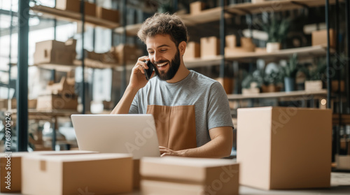 Cheerful small business owner multitasking with phone call and laptop amidst shipping boxes in modern e-commerce warehouse for efficient order fulfillment and customer service.