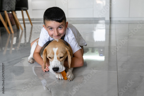 A young boy smiles while resting on a tiled floor beside his beagle. The dog focuses on chewing a tasty treat, showcasing their friendship in a cozy home setting.