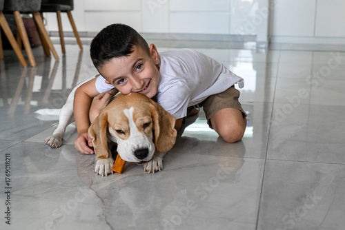 A cheerful boy hugs his beagle on a shiny floor, as the dog focuses on chewing a tasty treat. This scene captures a warm moment between a playful pet and a happy child.