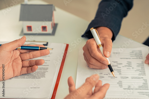 Signing a contract next to a model home, symbolizing home ownership. A real estate agent shows a small model home to a client with a calculator. Contract documents on a table.