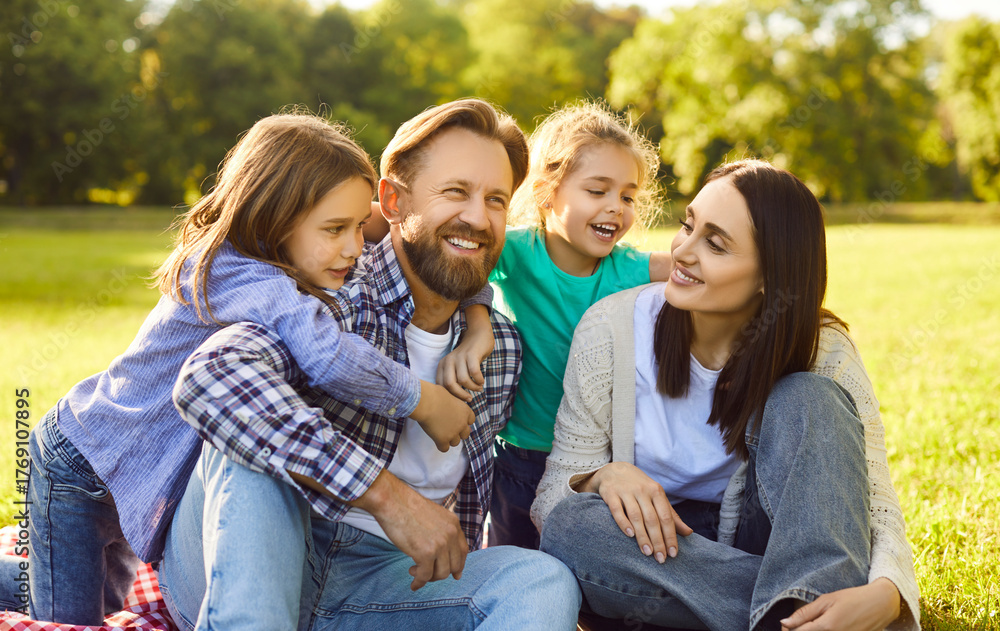 Fototapeta premium Young happy smiling family of four with son and daughter sitting on green grass in the summer park. Joyful parents enjoying sunny day in nature together with their kids. Family leisure concept.