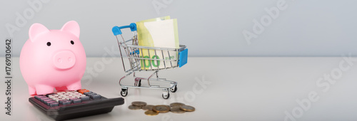 A pink piggy bank, calculator, and miniature shopping cart filled with money.