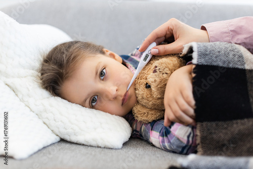A young girl lies on a couch with a thermometer in her mouth, being cared for by a parent. She is wrapped in a blanket with a teddy bear.
