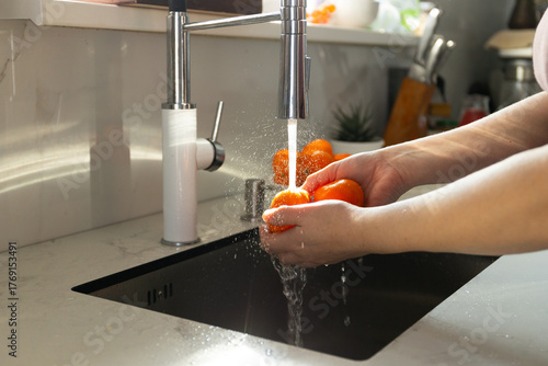 Fotografija Hands hold fresh oranges under running water at a clean kitchen sink, demonstrating the proper way to wash produce for hygiene before eating