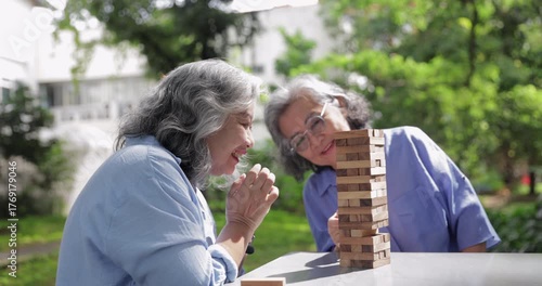 Two elderly women concentrate while playing a wooden block stacking game outdoors. Their serious expressions reflect focus, strategy, and mental engagement in a relaxing natural setting.