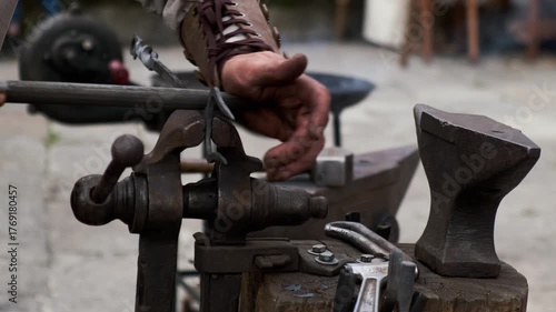 Static lateral close-up of a blacksmith smoothing a clamped metal piece with a hand file; anvil and hammer in soft background, assorted tools around the workbench.