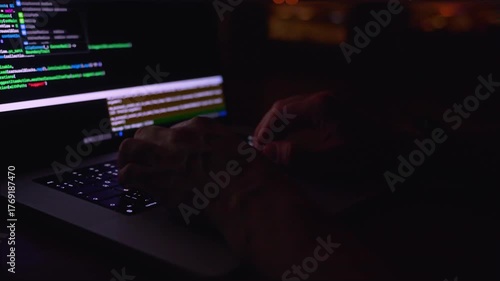 A young male programmer in front of a window with a lights of night city, on his laptop screen is code. out of focus. 