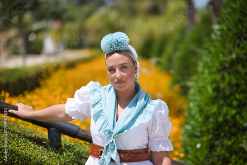 Foto Portrait of young, beautiful, blonde woman, with typical Andalusian flamenco costume to go on pilgrimage in white color with light blue shawl and flower, leaning on a railing in an outdoor park