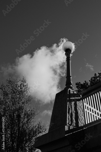 A dramatic black and white shot of a classic street lamp against a dark sky with a bright cloud. The vintage lamppost stands on an old stone bridge, creating a sense of timeless elegance