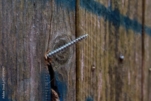 A silver self-tapping screw is partially screwed into a cracked, weathered wooden plank. The screw is at an angle, highlighting the texture and damage of the old wood.