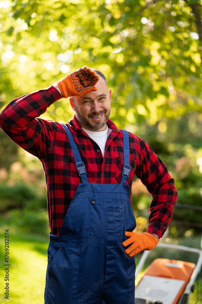 Obraz premium Professional gardener with protective gloves and goggles posing beside lawn cutting machine