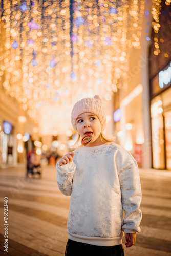 Young girl enjoying a lollipop in a festive shopping street during the evening hours