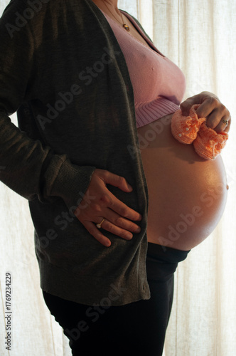 Portrait of a Pregnant Woman Standing in front of a Window Holding Small Shoes for Her Future Baby. Pregnancy Shooting in Home Interior