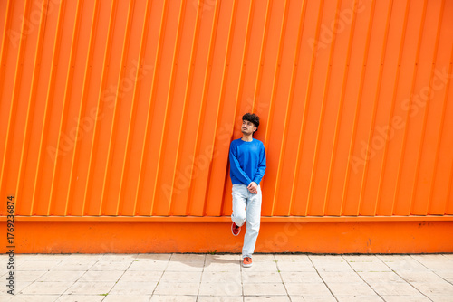Young man in blue sweater leaning against an orange wall outdoors
