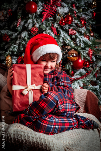 Happy little girl child in pajamas and festive santa hat smiling while sitting near christmas tree with gift box. Magic. New Year's eve. St. Nicholas Day. Present. Time of holidays and miracles.	