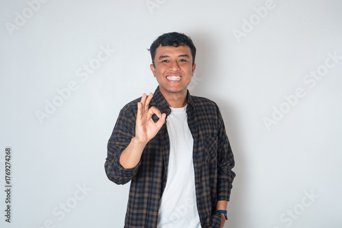 Smiling young Asian man in casual shirt doing ok sign with fingers isolated on white background
