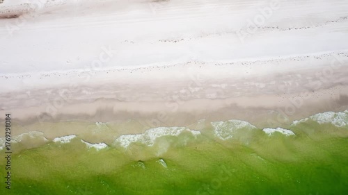 Emerald-green water rolls gently across the shore, creating soft ripples and foam patterns as it meets the pale white sand in a serene coastal scene.