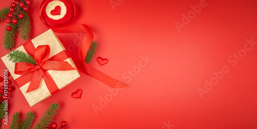 Christmas background with golden gift box and red ribbon on red backdrop, fir branches, berries and heart decorations, festive minimal composition with copy space for text, top view flat lay