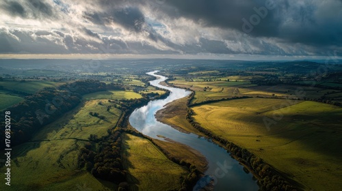 Aerial view of a meandering river flowing through a picturesque valley, with dramatic clouds casting shadows on the verdant landscape