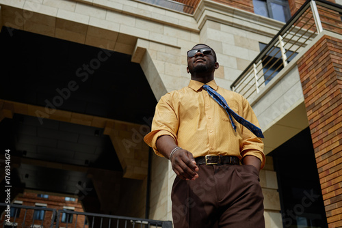 Young adult Black man walking confidently outdoors in urban setting, wearing sunglasses and patterned blue necktie, looking away from camera, modern architecture in background