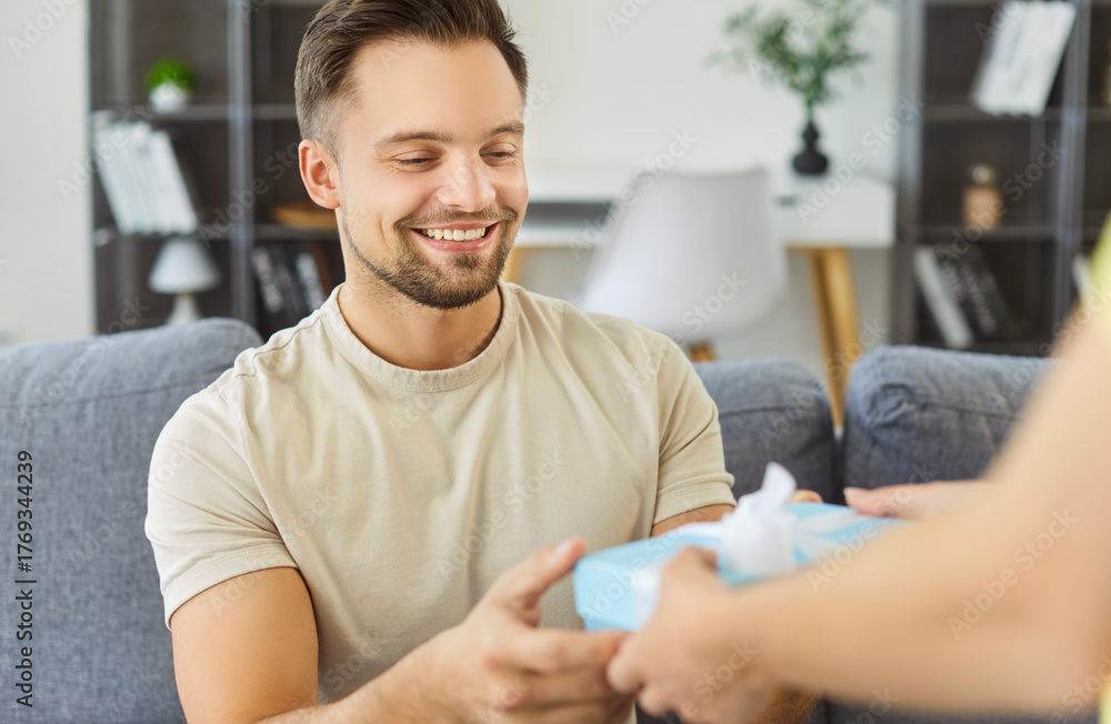 Fototapeta premium Happy man sitting on sofa receiving gift from hands offering, guy smiling warmly, happiness and gratitude, gift-giving for special occasion day, creating a joyful birthday, event party to share 