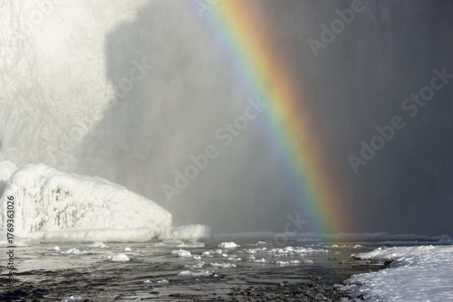 View of a vibrant rainbow arcing through the misty air, contrasting with the stark white of snow and ice-covered rocks, a scene of natural beauty, Skeioflotur, Rangárping eystra, Iceland.