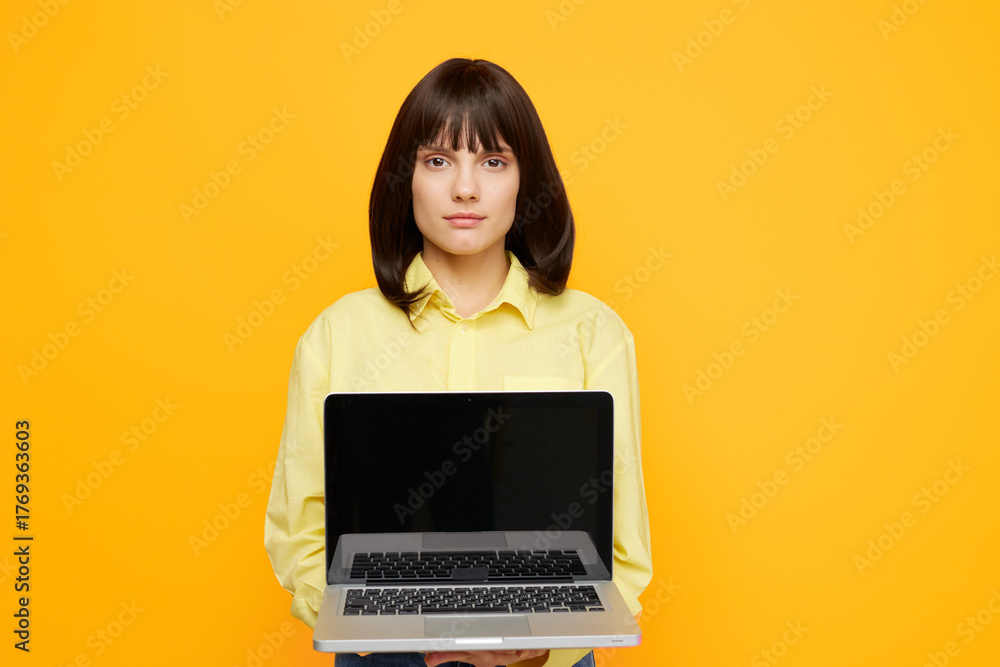 Fototapeta premium A calm woman stands against a bright yellow backdrop, holding an open laptop toward the camera. The image conveys focus, remote work, and digital creativity.