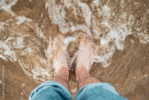 Canvas Print Caucasian male bare feet and pulled up jeans, standing on a sandy beach with shallow tidal waves sea water reaching the shore